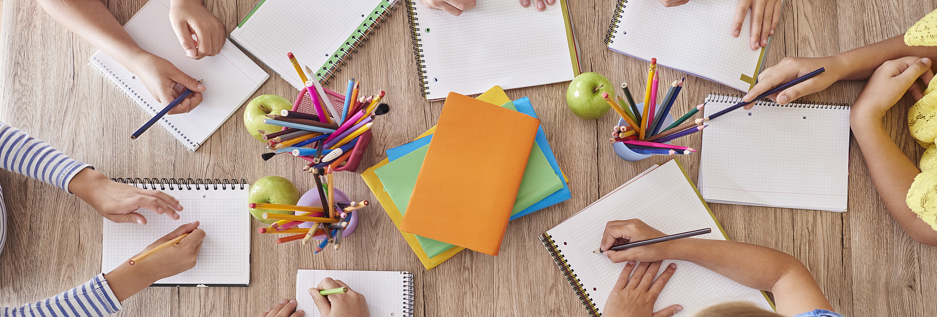 7 Schulkinder sitzen mit leeren Schreibblöcken und Stiften in der Hand um einen Schreibtisch herum
