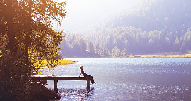 Eine Frau sitzt bei Sonnenschein am Ende eines Steges an einem See