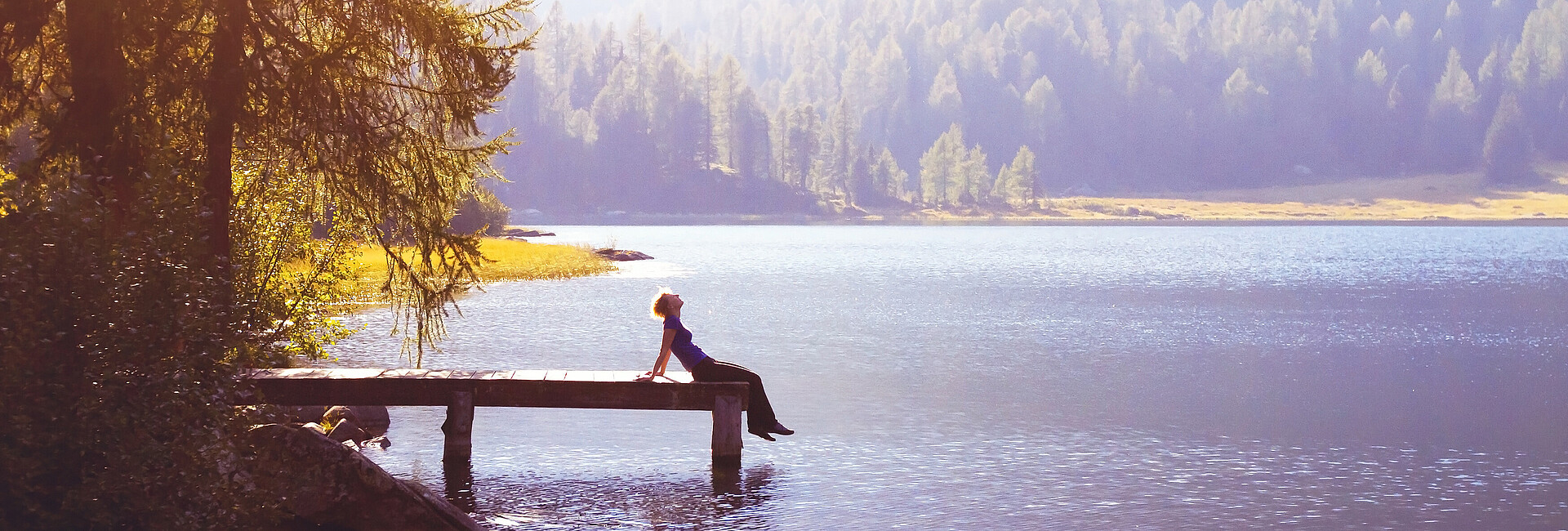 Eine Frau sitzt bei Sonnenschein am Ende eines Steges an einem See