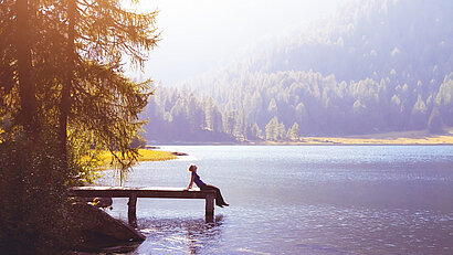 Eine Frau sitzt bei Sonnenschein am Ende eines Steges an einem See