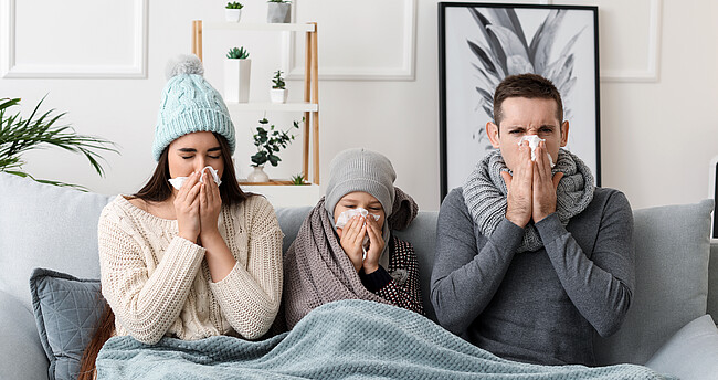 Erkältete Familie sitzt auf einer Couch unter einer Decke und putzt sich die Nase