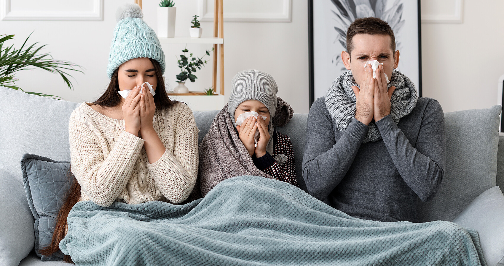 Erkältete Familie sitzt auf einer Couch unter einer Decke und putzt sich die Nase