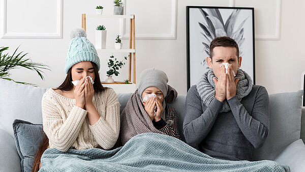 Erkältete Familie sitzt auf einer Couch unter einer Decke und putzt sich die Nase
