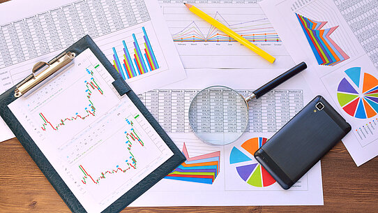 Diagramme, Bleistift, Lupe und ein Mobiltelefon liegen auf einem Tisch