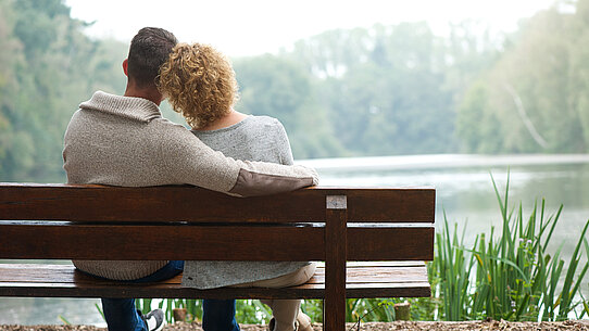 Mann hält Frau im Arm auf einer Bank an einem See