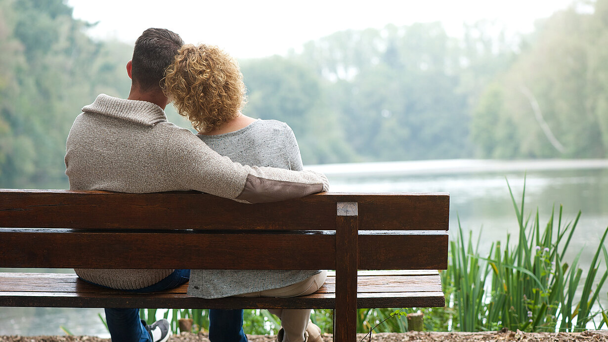 Mann hält Frau im Arm auf einer Bank an einem See