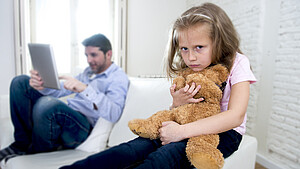 Trauriges Kind mit Teddybär, im Hintergrund Vater mit Tablet