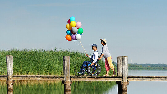 Mann im Rollstuhl hält eine Anzahl Luftballons, während er von einer Frau über einen Steg geschoben wird