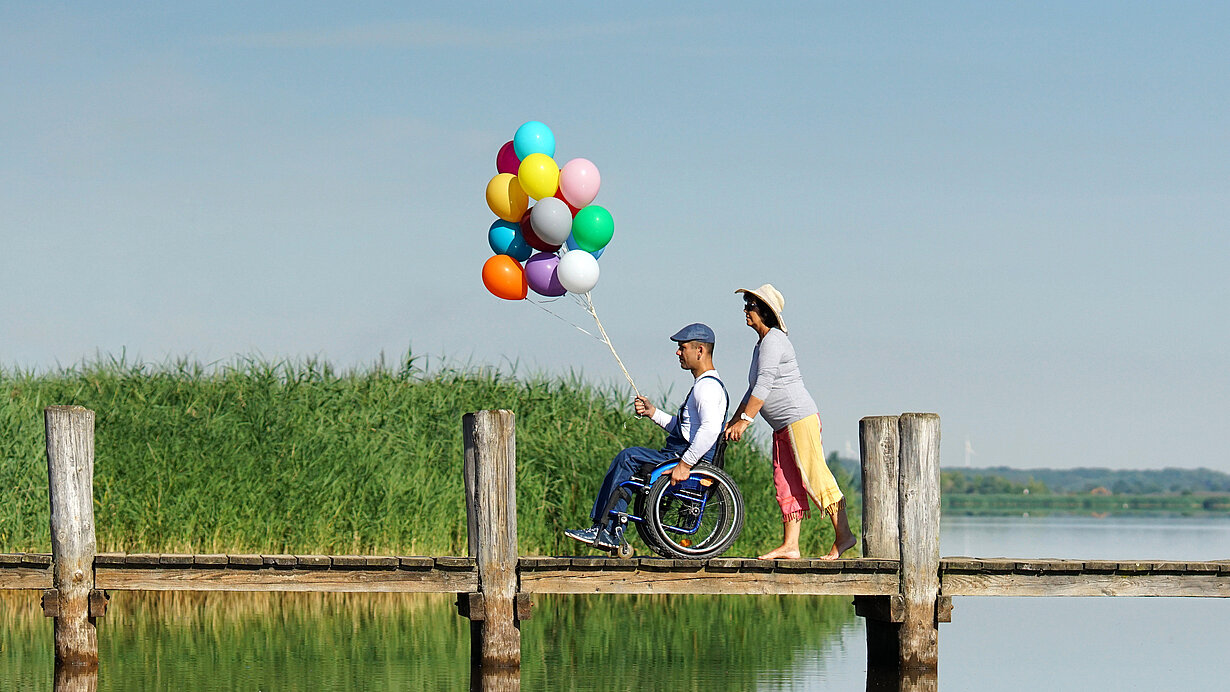 Mann im Rollstuhl hält eine Anzahl Luftballons, während er von einer Frau über einen Steg geschoben wird