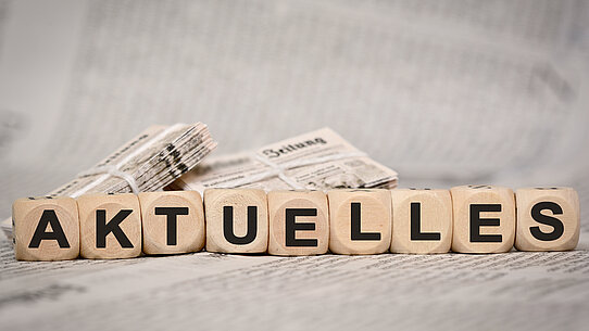9 Würfel vor einem Zeitungsstapel bilden das Wort "Aktuelles"
